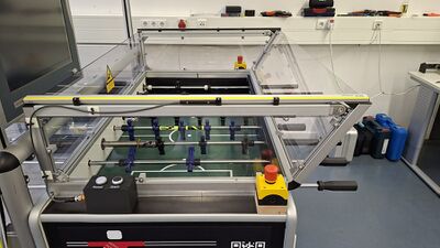 Der Kickertisch soll von oben ohne Reflexionen durch Plexiglasplatten einsehbar sein. Gleichzeitig muss die Gefahr gebannt werden, sich die Finger quetschen zu können. Deswegen wurde in sicherer Höhe über dem Spielfeld ein Lichtgitter installiert. So haben die Spielfiguren Zeit, um zum Stillstand zu kommen, sollte doch einmal jemand während des Spielbetriebs auf das Spielfeld greifen. Mit dem weißen und dem schwarzen Taster vorn kann die Maschine nach Unterbrechung des Lichtgitters vom Bedienpersonal wieder in Gang gesetzt werden.